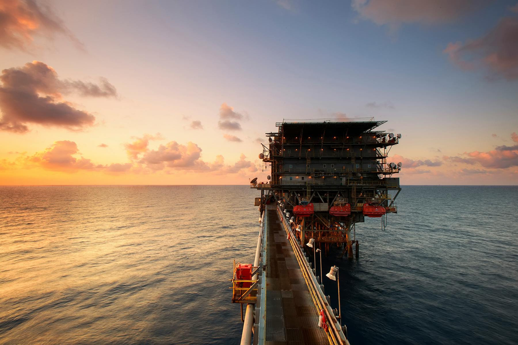 An offshore oil platform set against a sunset sky, surrounded by calm ocean waters.